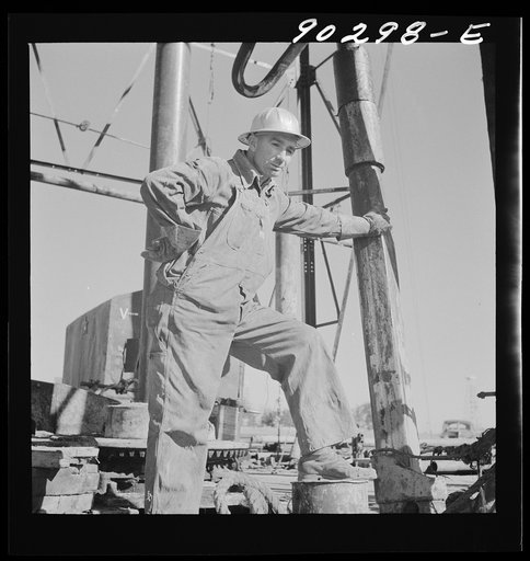 J.E. Schenermann, driller from Oklahoma, but worked in the Kansas oil fields about ten years; on pile of drill pipes near oil well in Goodrich field of Continental oil company. Valley Center oil field near Wichita, Kansas