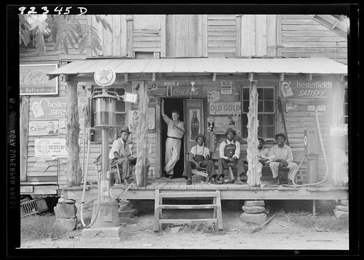 Untitled photo, possibly related to: Country store on dirt road. Sunday afternoon. Note the kerosene pump on the right and the gasoline pump on the left. Rough, unfinished timber posts have been used as supports for porch roof. Negro men are sitting on the porch. Brother of store owner stands in doo