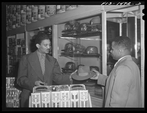 Mr. Claude Walker trying on a new hat in the Henry C. Taylor Store for Men. The salesman is Mr. William Strong, who is manager of the store. 47th Street near State Street, Chicago, Illinois