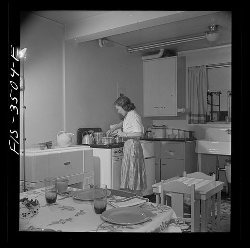 Greenbelt, Maryland. Federal housing project. Mrs. Leslie Atkins preparing dinner in her kitchen, one end of which is the dining room. Notice the mangle and washing machine on either side of the stove