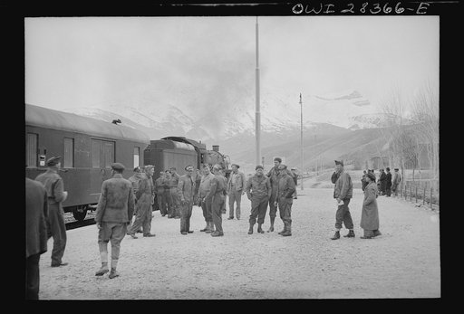American and British soldier trainmen standing about at a station on the route for supplies to Russia. An American engine is seen at the head of the train at left, somewhere in Iran