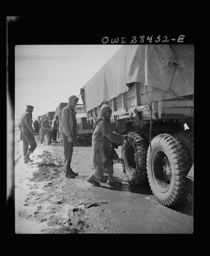 Somewhere in the Persian corridor, a United States Army truck convoy carrying supplies for the aid of Russia. An Iranian boy giving the American driver a helping hand at putting on chains before the trucks go into a snow bound pass through the mountains