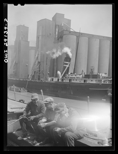 Untitled photo, possibly related to: Buffalo, New York. Grain boat from Duluth unloading wheat at a grain elevator on the Erie Canal. Men sit on the edge of an open hatch to guide the ropes of the shovel which scoops the grain into the hatch where it can be drawn by the "leg" into the tower on an en