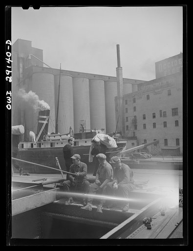 Untitled photo, possibly related to: Buffalo, New York. Grain boat from Duluth unloading wheat at a grain elevator on the Erie Canal. Men sit on the edge of an open hatch to guide the ropes of the shovel which scoops the grain into the hatch where it can be drawn by the "leg" into the tower on an en