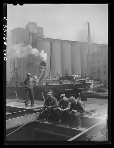 Buffalo, New York. Grain boat from Duluth unloading wheat at a grain elevator on the Erie Canal. Men sit on the edge of an open hatch to guide the ropes of the shovel which scoops the grain into the hatch where it can be drawn by the "leg" into the tower on an endless belt of buckets