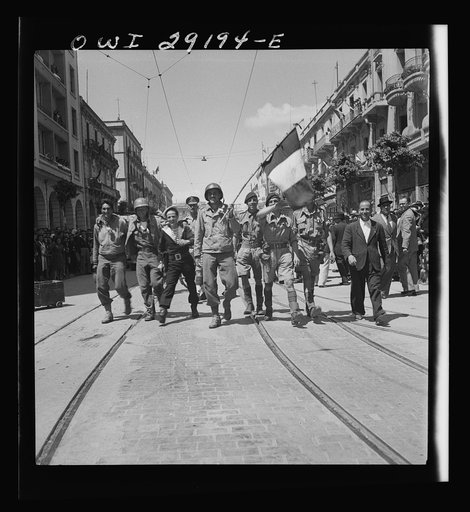 An American soldier, carrying a tricolor, leads a happy group of Allied sailors and soldiers down the middle of a main thoroughfare in Tunis