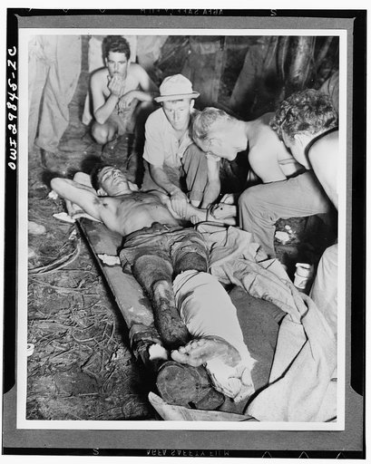 A wounded American soldier gets a blood transfusion while assistants hover about the improvised cot. The scene is "somewhere in New Guinea"