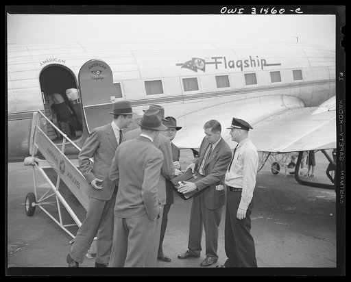 An officer and a government official (?) meeting two men at the American Airlines "Flagship" passenger plane and examining the contents of a brief case, in a scene in the motion picture "Black Marketing," produced by the U.S. Office of War Information (OWI) and released by August 19, 1943