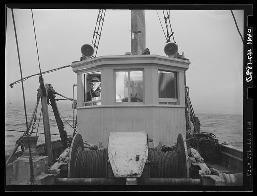 Gloucester, Massachusetts. With the light from the compass binnacle illuminating his face, Captain John Riberia [i.e., Ribeiro] guides "Old Glory" back to the port. Decks have been cleared and fish stored in ice until return to port