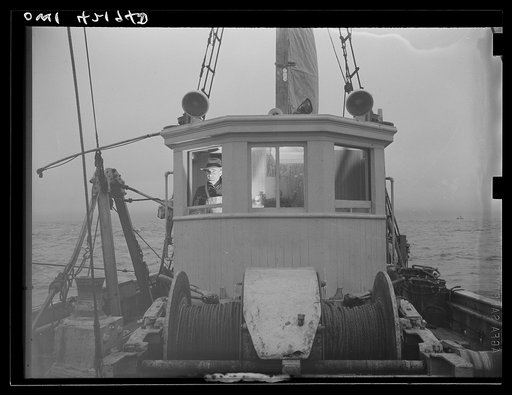 Gloucester, Massachusetts. With the light from the compass binnacle illuminating his face, Captain John Riberia [i.e., Ribeiro] guides "Old Glory" back to port. Decks have been cleared and fish stored in ice until return to port