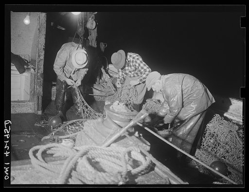 On board a fishing vessel out from Gloucester, Massachusetts. Fishermen repairing the nets. In the foreground is a trawl cable around the bollard