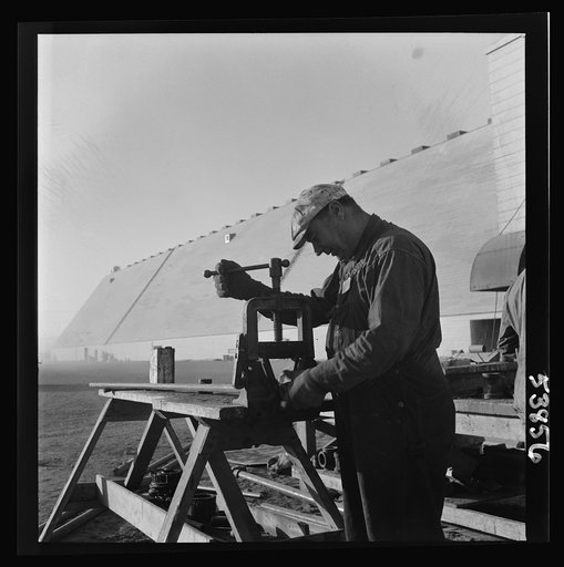 Las Vegas, Nevada. A pipe fitter at work at a pipe, or machinist's vise, near one of the 540-foot peat storage buildings at the Basic Magnesium Incorporated plant in the southern Nevada desert