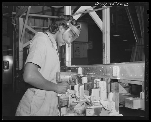 A Negro woman at North American Aviation uses an electric hand drill to drill holes in a sheet metal assembly prior to riveting