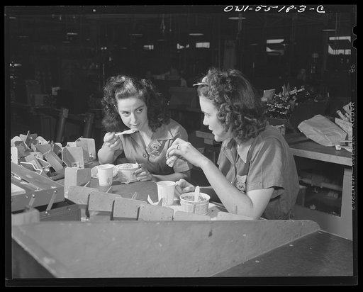 Girls eat lunch together at their work benches. Lunch carts (Travelunch) go through the factory and enable employees of North American Aviation to buy their lunches without leaving the plant