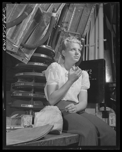 Surrounded by sub-assemblies for the North American B-25 bombers and P-51 fighters, this woman employee on the "swing shift" enjoys her lunch at the bench