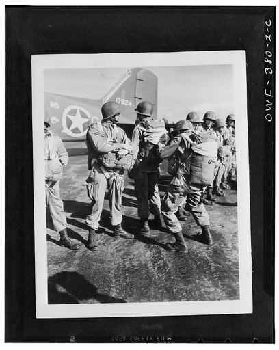 U.S. paratroopers inspecting each others' parachutes before taking off on maneuvers somewhere in England. Note the camouflaged helmets