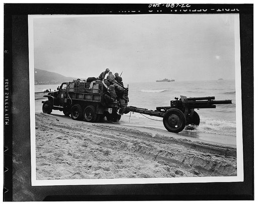 Two-and-a-half ton prime mover towing a 105 millimeter howitzer along the Algerian beach. Troop ships can be seen in the background