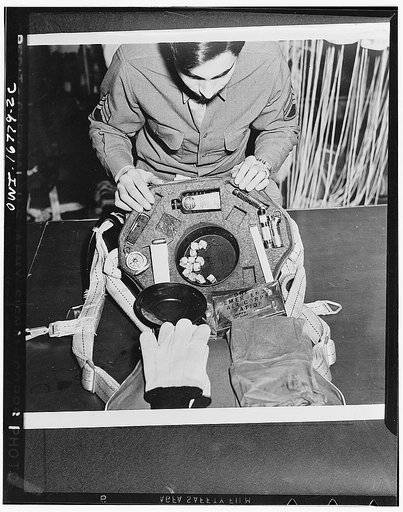 Sergeant Tony Gaudiello, of Uniontown, Pennsylvania, somewhere in the European war theater, inspecting the contents of the kit which fits into the back pad of a parachute. It contains, among other things, concentrated food, emergency ration, a frying pan, knife, lighter, compass, bullets, gloves, an