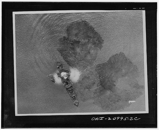 A 7,000 ton Japanese merchant vessel after a direct hit by India Air Task forces near Rangoon, India. A bomb is dropping in the lower right part of the picture and small boats are trying to carry the crew members away from the sinking vessel. The waters are covered with oil slick