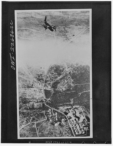 Operations in North Africa by Allied air forces. A light bomber of the South African air force comes in to bomb a large enemy barracks in Mareth village. Low clouds are above the target area. In the right foreground, bombs dropped by the previous aircraft can be seen exploding on the target