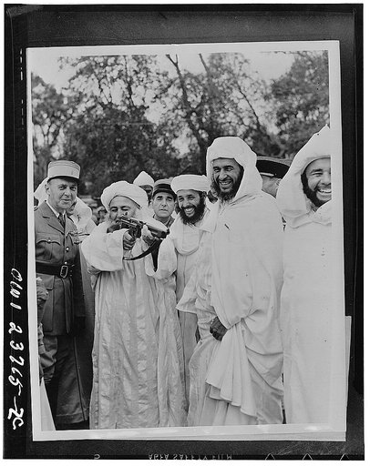 Pashas, caids and French officers looking on as His Excellency, the Pasha Ali draws a bead with a tommy gun. Pasha Ali, now 75, is the leader of one of the last of the Berber tribes to submit to the French occupancy of  Morocco in the early part of the 20th century