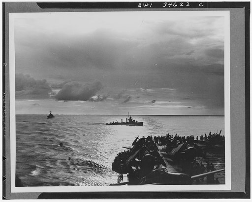 A watchful U.S. destroyer cuts through the wake of a U.S. aircraft carrrier on patrol as a setting sun brings to a close the first day of fighting off North Africa