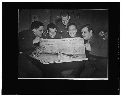 Men from New York State crowding around an out-dated copy of a New York newspaper at an American Red Cross club somewhere in England. Left to right: Private First Class Ted Rothstein, Bronx; Private Gene Binchoni, Bronx; Sergeant T. David Bernstein, Brooklyn; Private Jack Perino, Bronx, and Private 