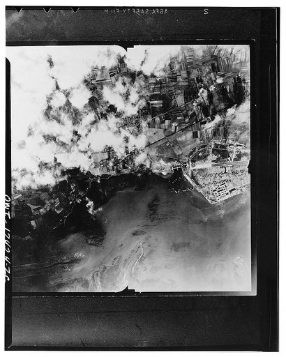 Cagliari, Sardinia. 1942(?) The airdrome billowing smoke and flames following an attack by US Army Air forces. Fires are eating at the administration buildings at a corner of the base and others are raging at the runway. At another point of the airdrome an ammunition dump has been hit. This was the 