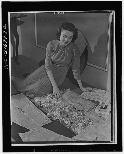 Wartime conservation through home sewing is demonstrated by Powers model Cynthia Hope who works with a fabric-saving dress pattern. All patterns are now adjusted to WPB (War Production Board) conservation rules. Average saving: 1/8 yard per dress. Cynthia cuts the top of her contrast dress with a re