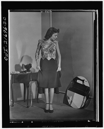 Dress almost finished, Cynthia adjusts her skirt length. The dress's hem will be no deeper than  WPB (War Production Board)-approved two inches. Cynthia's fabric-conserving pattern requires only three yards of 39-inch material. For all its fashionable appearance it wastes not one inch of material on