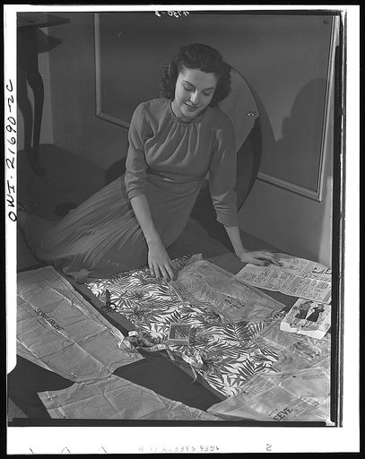 Wartime conservation through home sewing is demonstrated by Powers model Cynthia Hope who works with a fabric-saving dress pattern. All patterns are now adjusted to WPB (War Production Board) conservation rules. Average saving: 1/8 yard per dress. Cynthia cuts the top of her contrast dress with a re