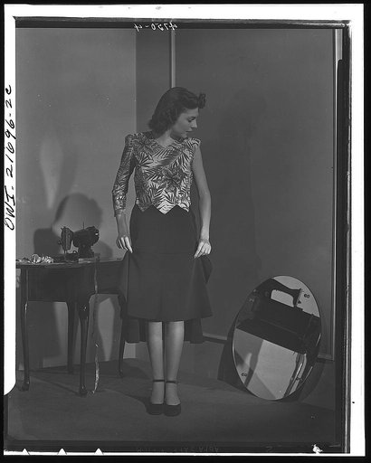 Dress almost finished, Cynthia adjusts her skirt length. The dress's hem will be no deeper than  WPB (War Production Board)-approved two inches. Cynthia's fabric-conserving pattern requires only three yards of 39-inch material. For all its fashionable appearance it wastes not one inch of material on