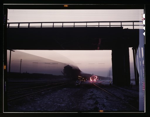 View in a departure yard at C & NW RR's [i.e. Chicago and North Western railroad's] Proviso (?) yard at twilight, Chicago, Ill. Brakeman is signaling with a red flare and the train is going by during exposure