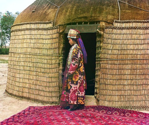 Full-length profile portrait of a woman, possibly Turkman or Kirgiz, standing on a carpet at the entrance to a yurt, dressed in traditional clothing and jewelry