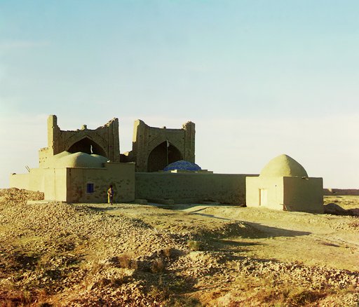 Walled adobe structure with domes and arches in desert area, with man posed in front