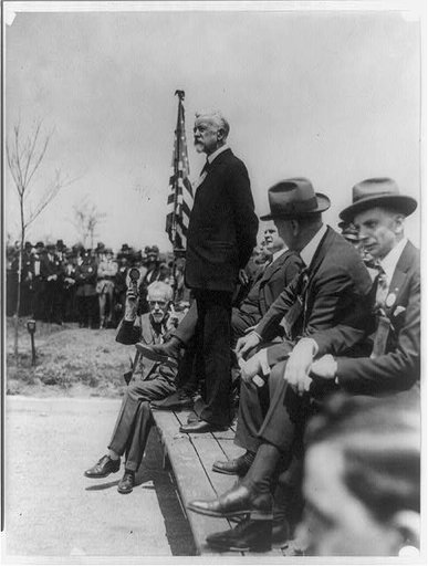 Henry Cabot Lodge standing on platform outdoors making a speech; man seated on platform with hearing aid or recording device