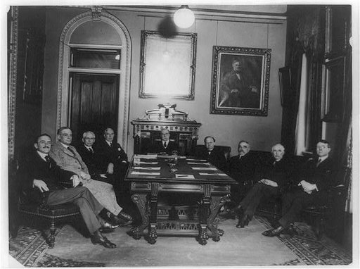 Charles Evans Hughes seated at head of table, with eight other men around the table in the Diplomatic Room of the State Department