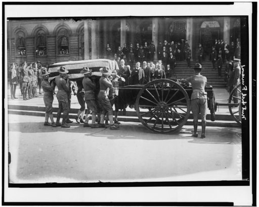 Soldiers loading flag-draped casket of former New York City mayor John Purroy Mitchel onto wagon, with Theodore Roosevelt and other officials holding their hats in respect