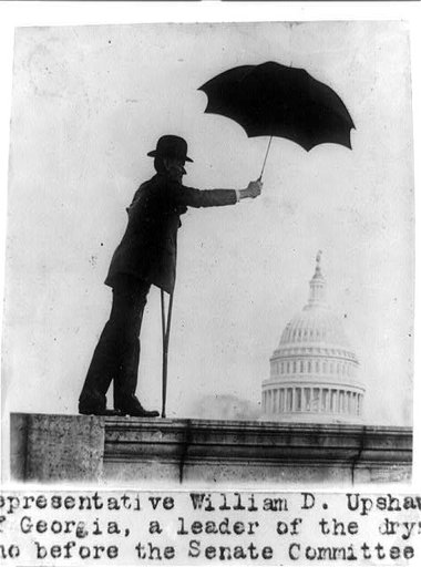 Representative William D. Upshaw of Georgia, a leader of the drys, standing on railing with crutch and holding umbrella, with dome of the U.S. Capitol under the umbrella
