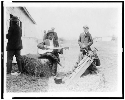 Two Afro-American men, outside of stable, with Kazoos in their mouths, one of them seated on haybale and playing guitar, the other playing homemade string instrument, Washington, D.C. or New York City