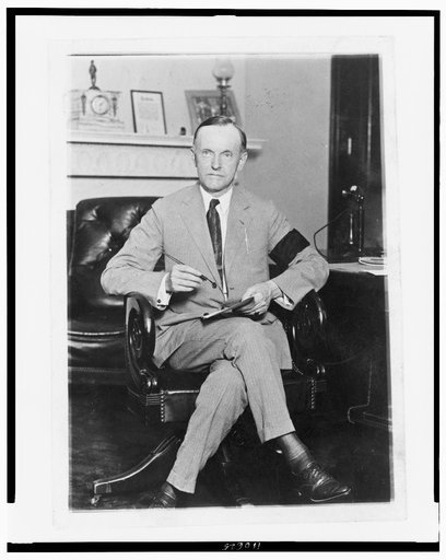 Calvin Coolidge, full-length portrait, seated at desk, facing front, holding pen and paper, wearing black armband in mourning for President Harding