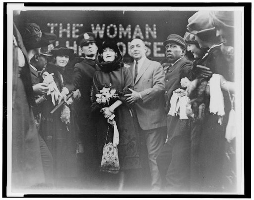 Mildred Harris Chaplin, full-length portrait, standing in crowd, facing front, guarded by a policeman