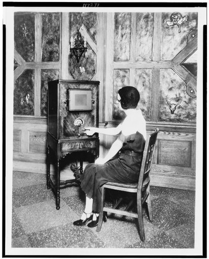 Woman, full-length, seated, facing left, turning dial of a console radio