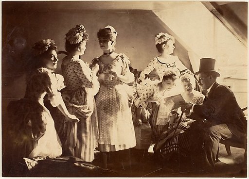 [A Group of Six Costumed Women Posed in Interior with Top Hatted Gentlemen]