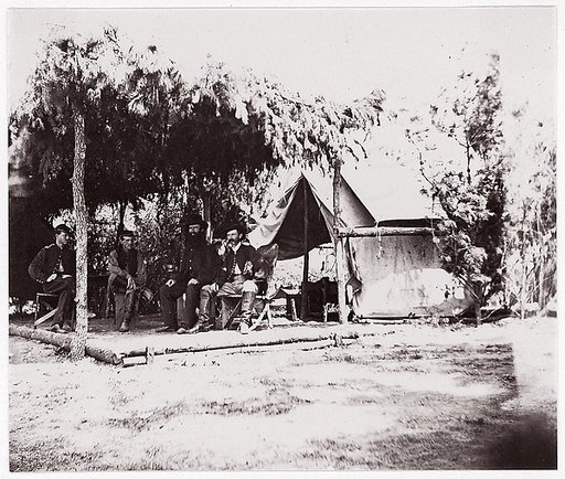 [Four men in camp under a lean-to of pine boughs]