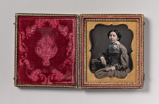 [Seated Young Woman Wearing Collar with Large Bow, Resting Arm on Table]
