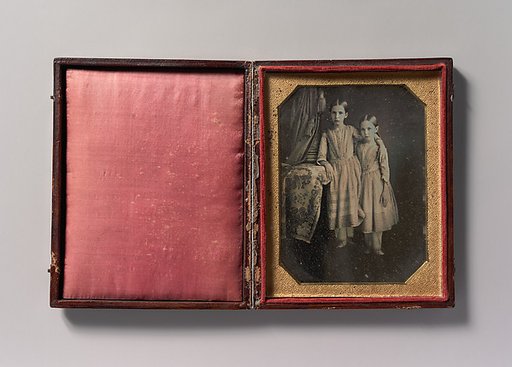 [Two Identically Dressed Young Girls Standing Next to a Table]