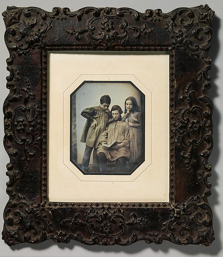[Three Children, One Seated Holding an Open Book, the Other Two Standing, in Front of a Light Background]