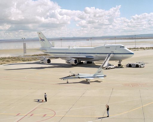 White clouds and a flooded lakebed form a backdrop as a T-38 support aircraft taxies across the ramp in front of NASA's Boeing 747 Shuttle Carrier Aircraft