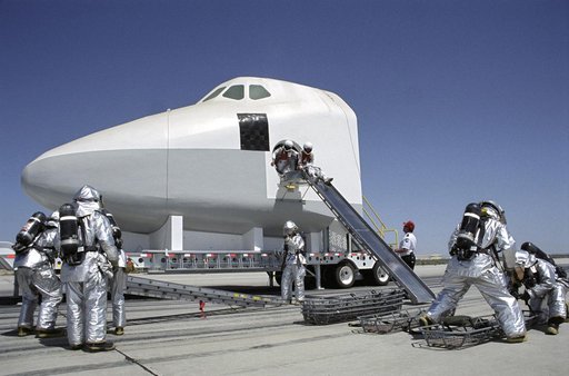 While rescue personnel prepare evacuation litters, two stand-in "astronauts" prepare to use an exit slide from a Shuttle mockup during a training exercise